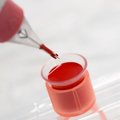 Close-up of a tattoo machine tip applying the Orange Coral - Tina Davies Lust Lip pigment into a small container, demonstrating its smooth consistency.